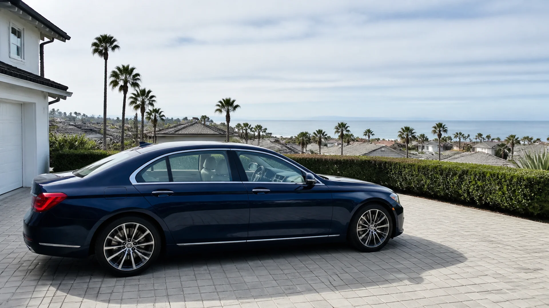 Blue sedan viewed from side on paved coastal driveway, with palms, rooftops and distant ocean horizon