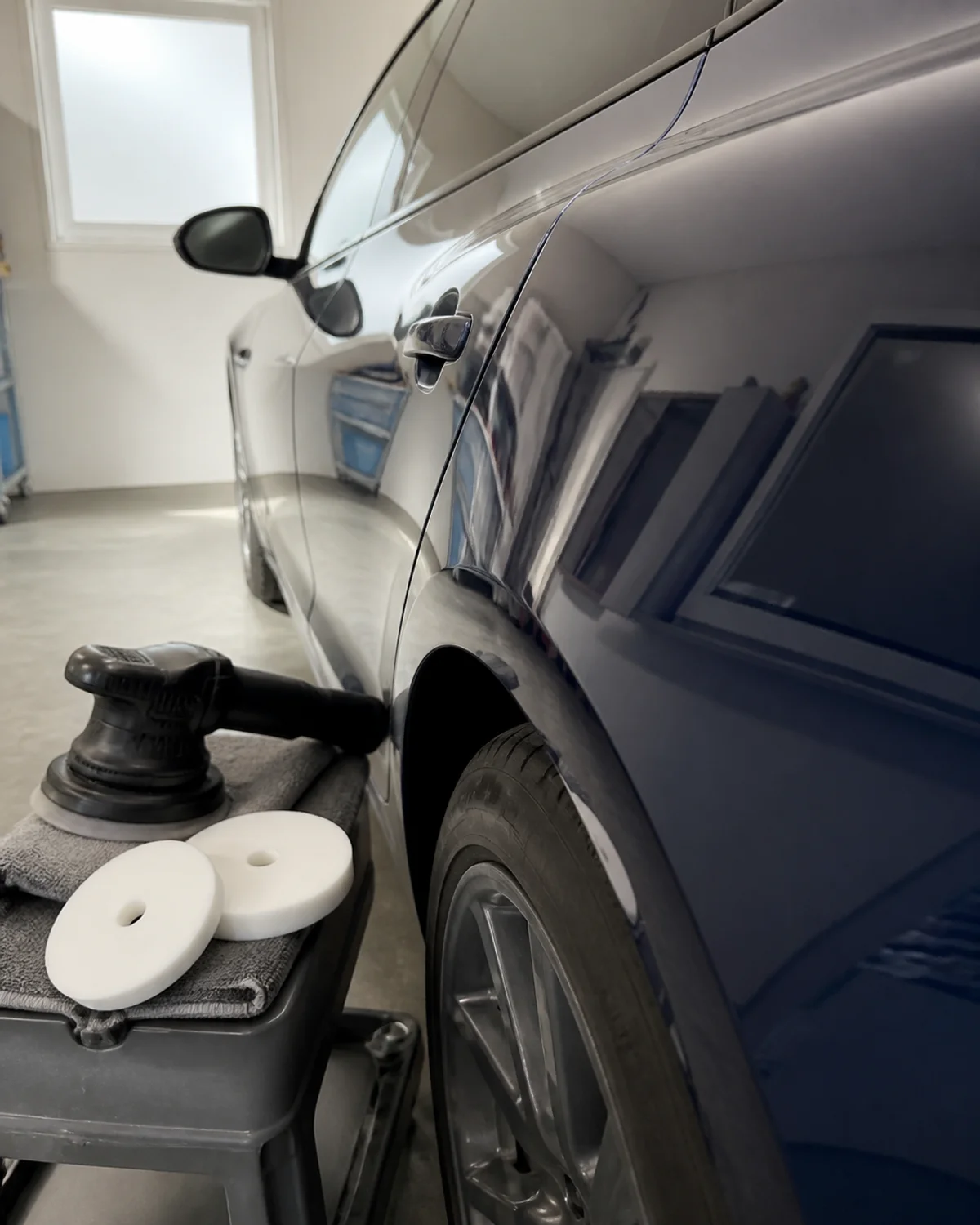 Glossy dark blue door and rear quarter panel reflecting garage walls beside a polisher, pads and towel